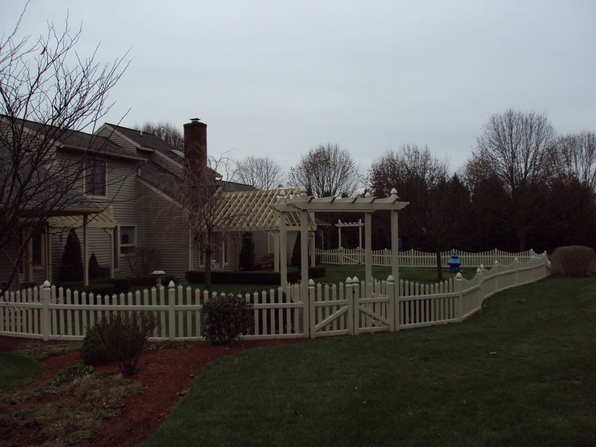 Cream picket fence surrounds a house with a pergola and trees in the background on an overcast day.
