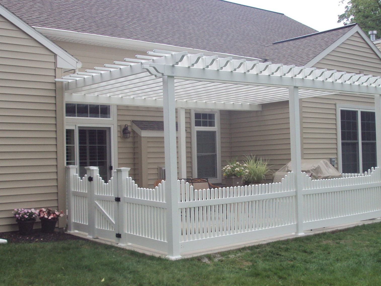White pergola and fence on a patio, attached to a beige house, with greenery and furniture inside.