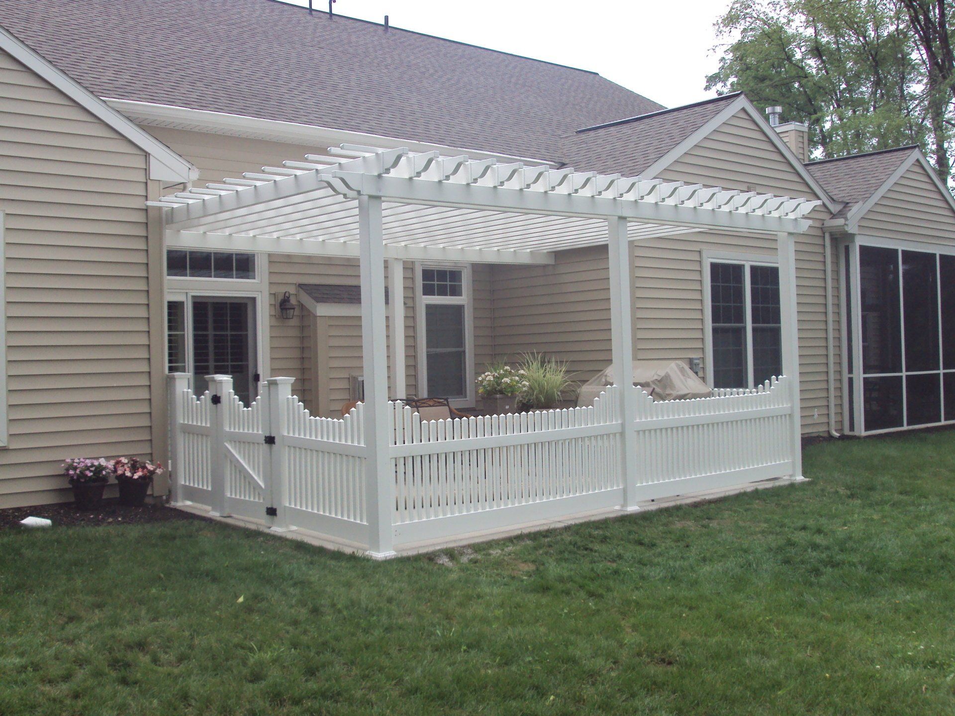 White pergola and picket fence on a patio attached to a beige house, with green grass in the foreground.