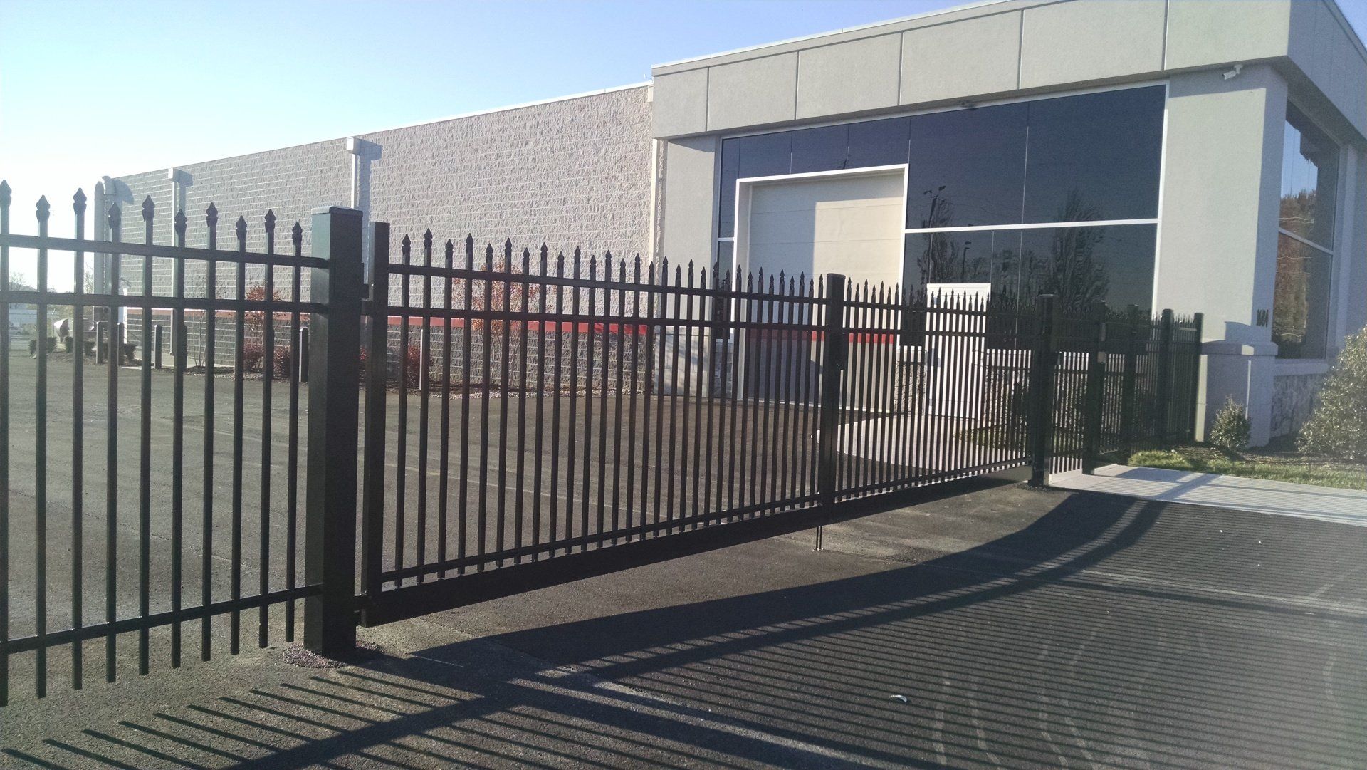 Black metal fence in front of a building with a large window and door. Sunny day, shadows on the ground.