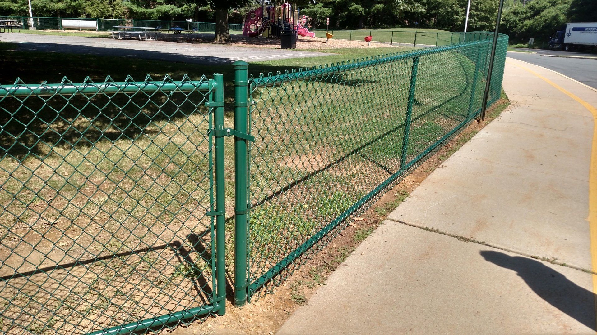 Green chain-link fence bordering a sidewalk and grassy area near a playground.