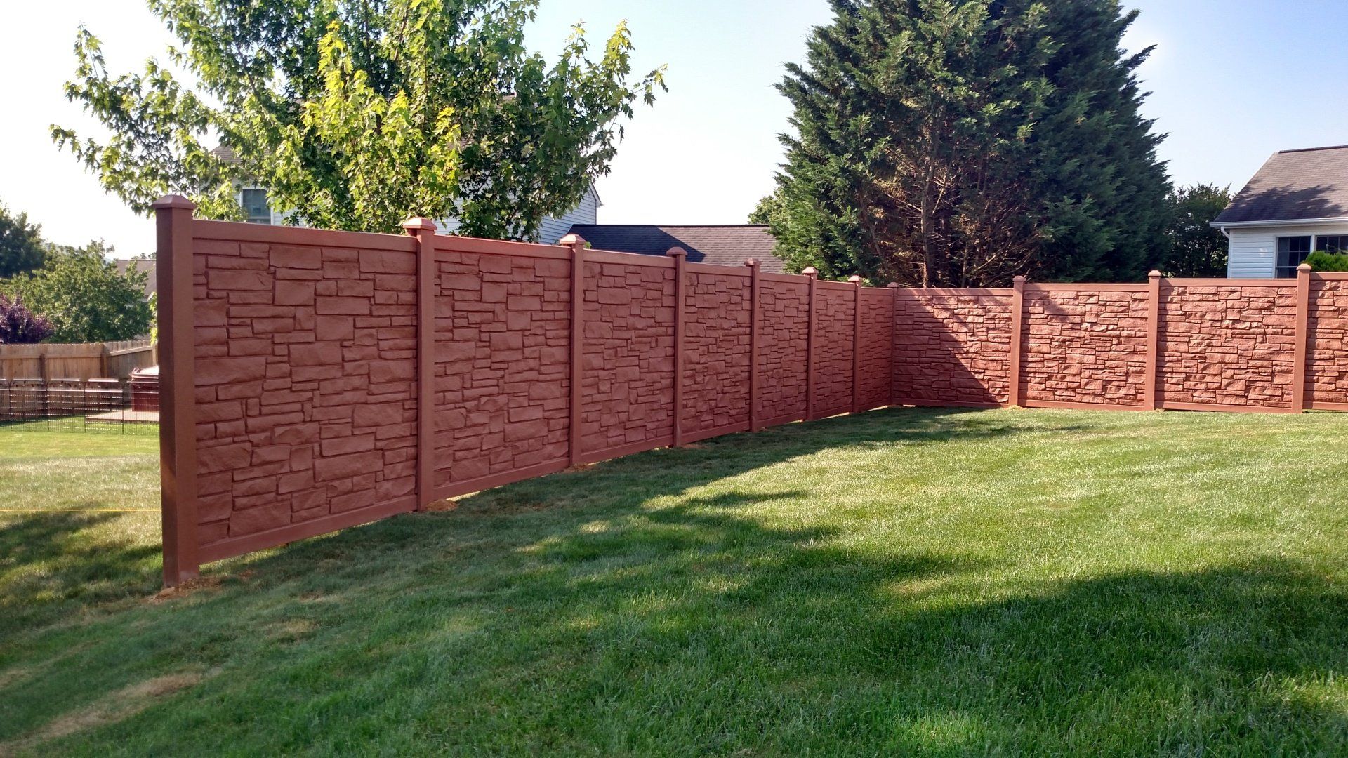 Brown textured fence in backyard with green grass and trees.
