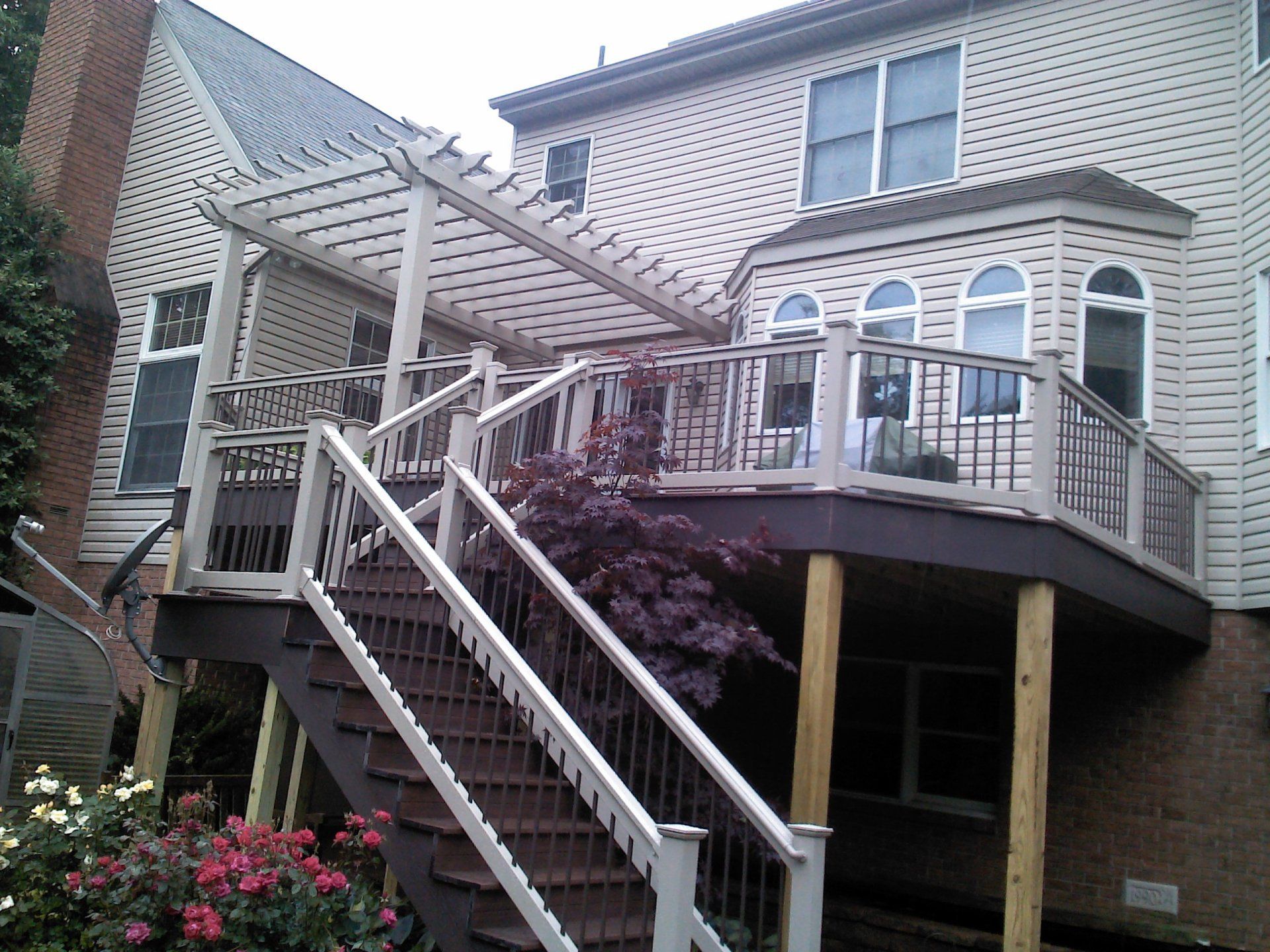 Multi-level wooden deck with pergola and stairs attached to a beige house, brown deck, and white railings.