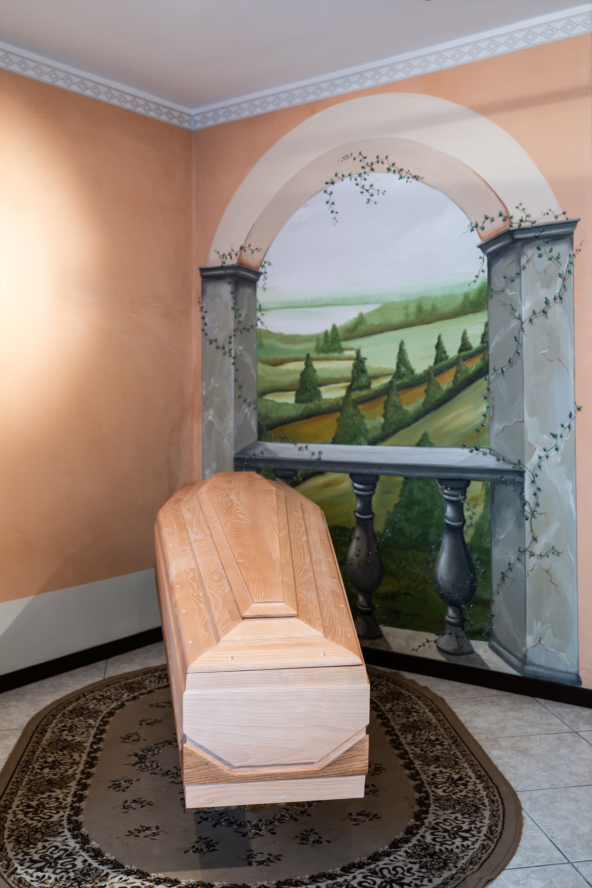 Una bara di legno è appoggiata su un tappeto davanti a un murale raffigurante un paesaggio.