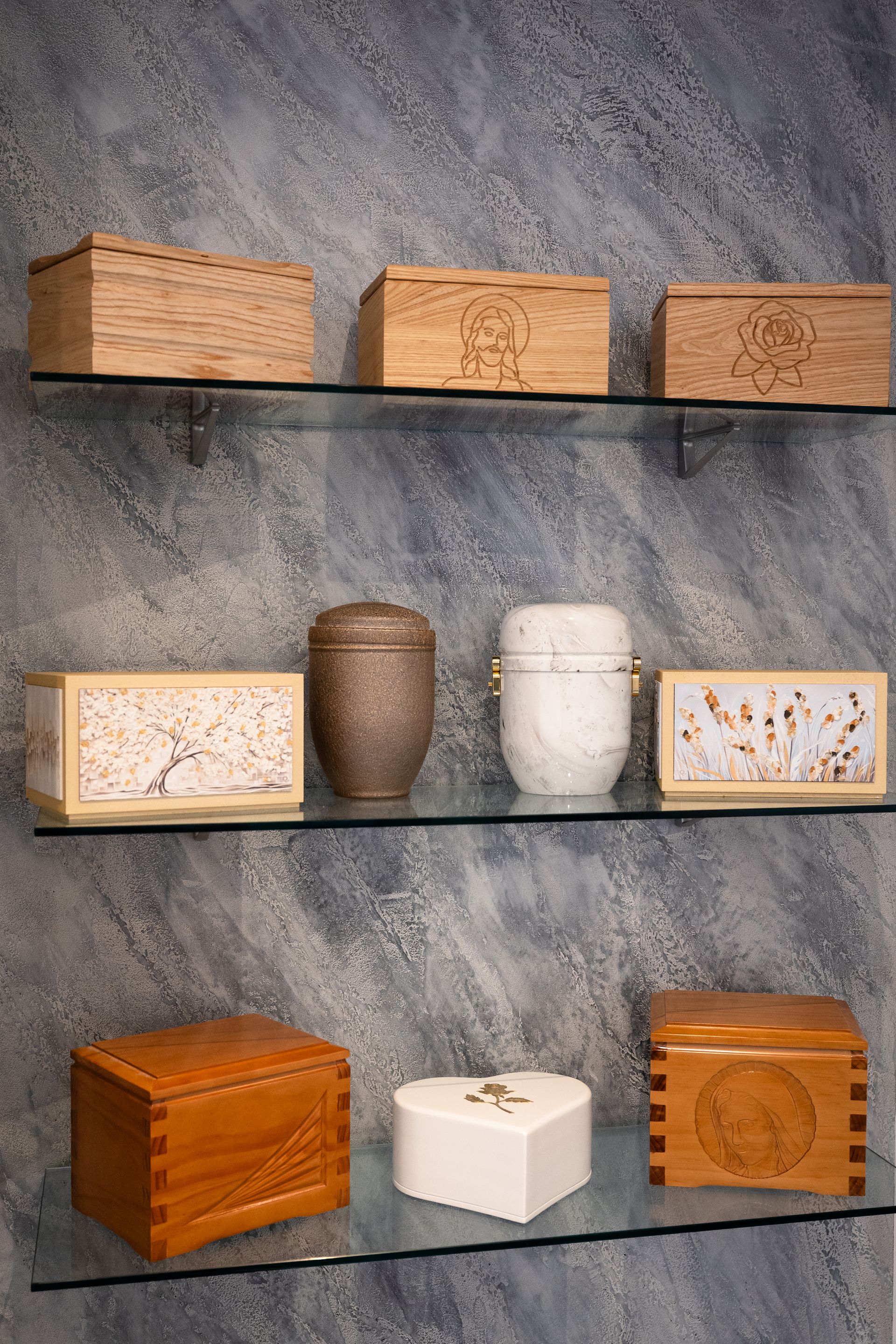 Urne decorative e in legno esposte su mensole di vetro contro una parete grigia testurizzata.