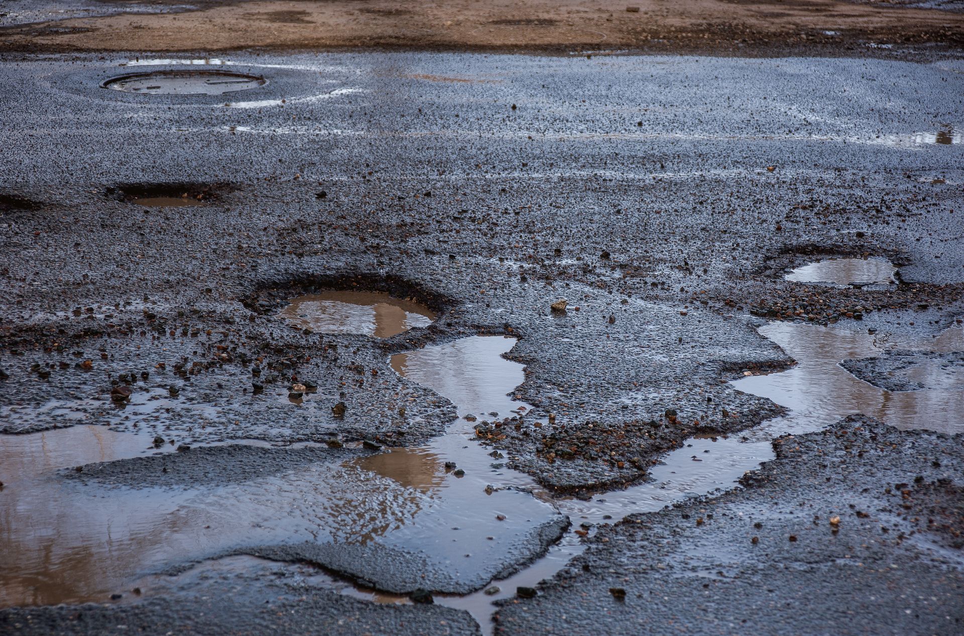 A damaged asphalt road with multiple potholes filled with rainwater, showing uneven surfaces and gravel.