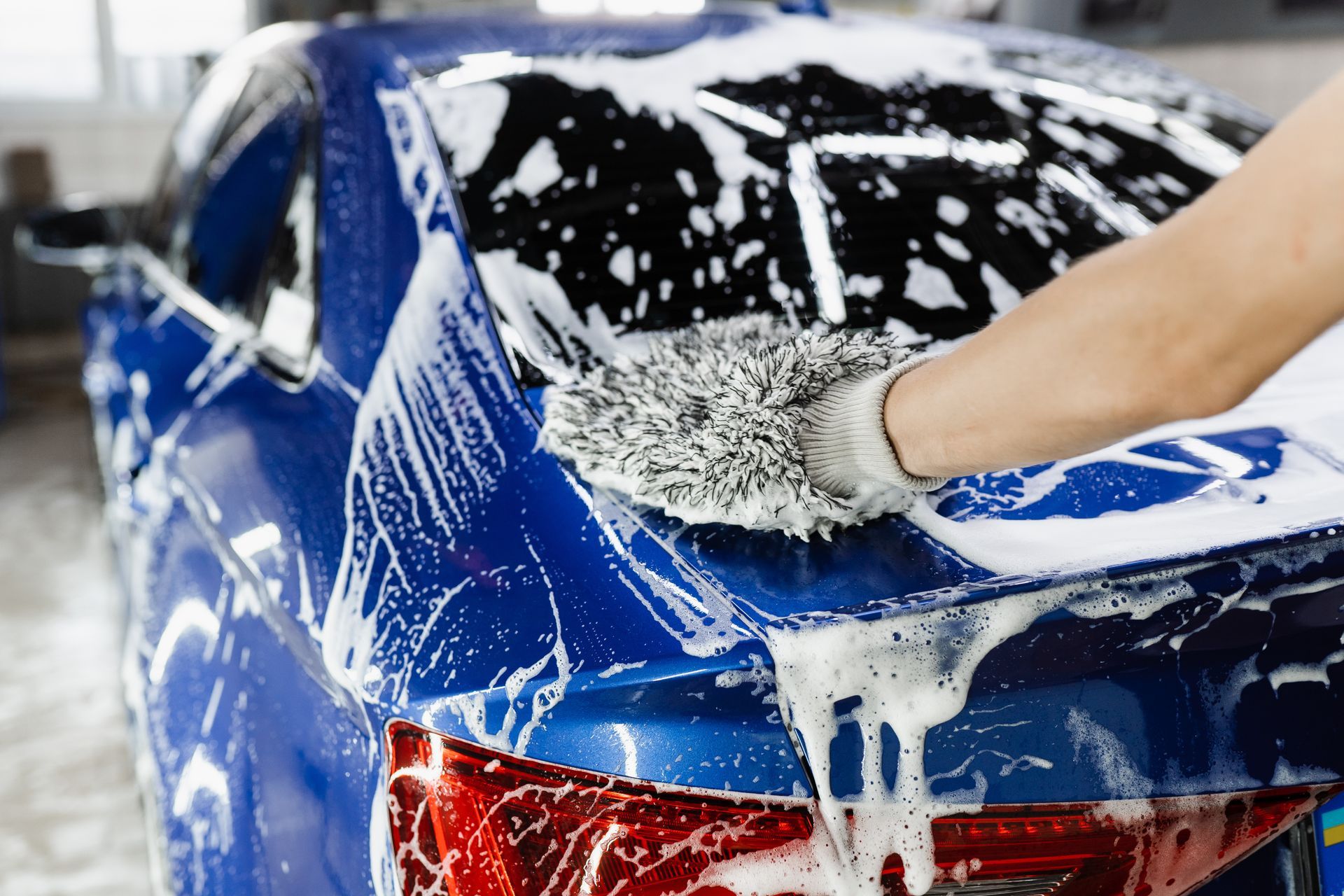 A hand using a fluffy wash mitt to clean soap suds off the trunk of a blue car.