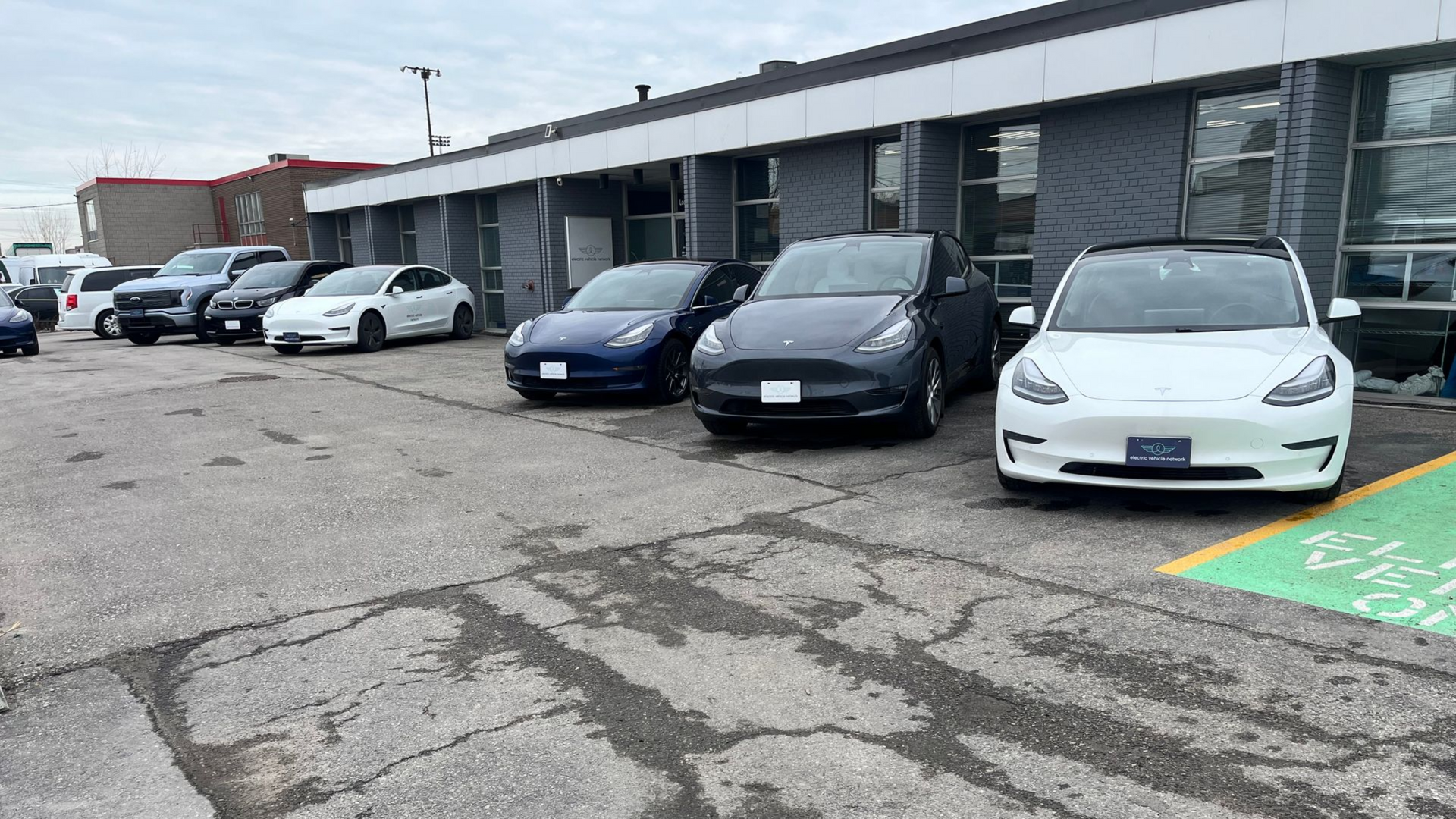 Cars parked outside a gray building. A line of Teslas, various colors, are in the foreground.