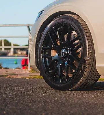 Black car wheel with black rim and tire against a waterfront backdrop.