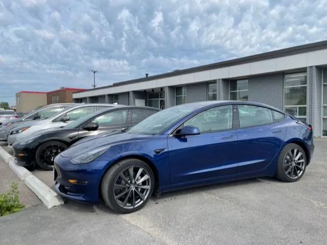 Blue Tesla sedan parked in front of a building. Other cars are visible nearby, cloudy sky.