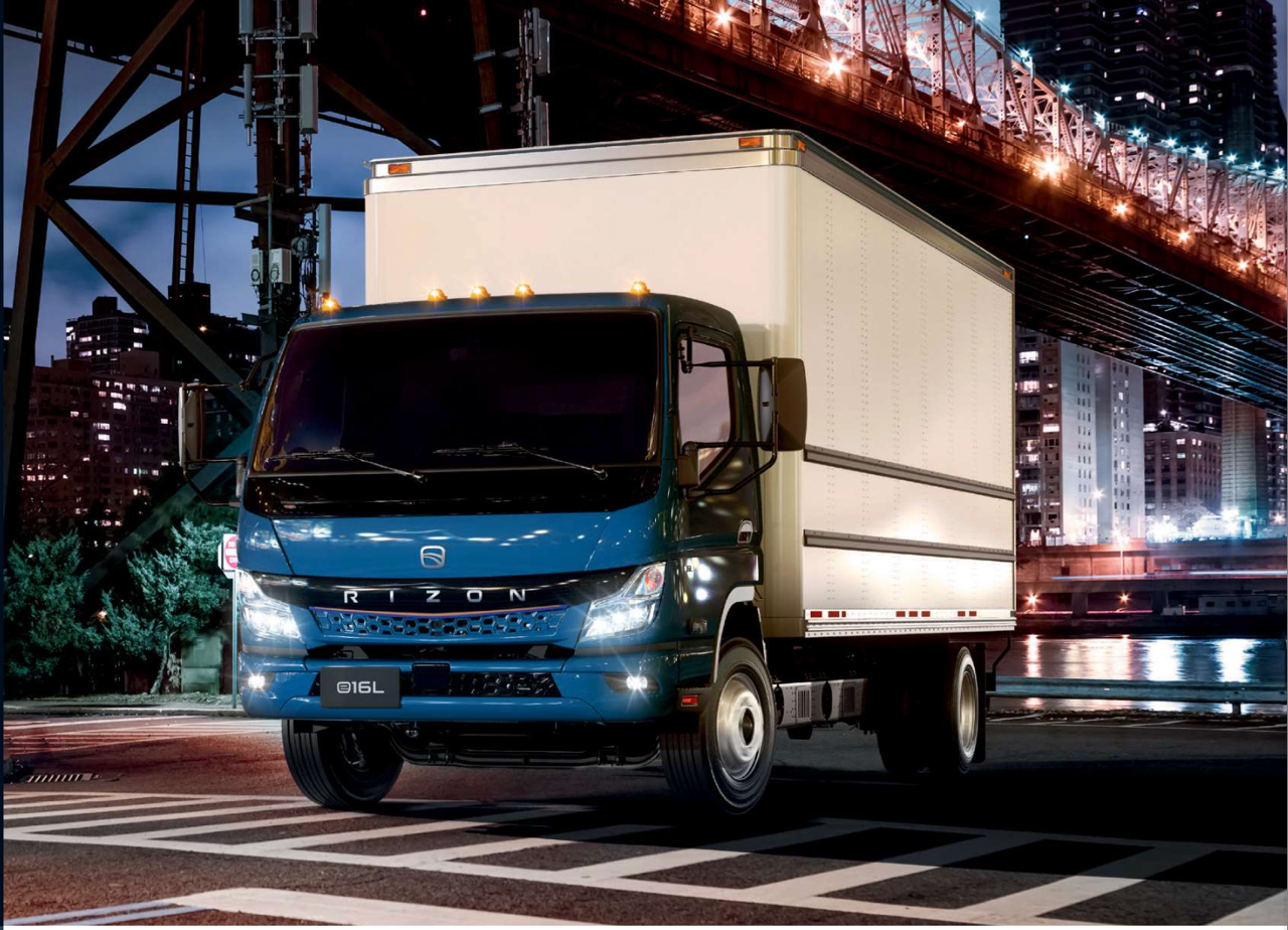 A blue box truck parked on a city street at night under a bridge with illuminated city buildings in the background.