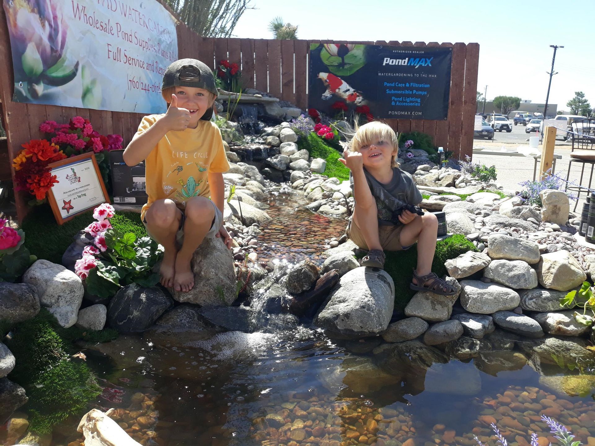 Two young boys are sitting in front of a pond and giving a thumbs up