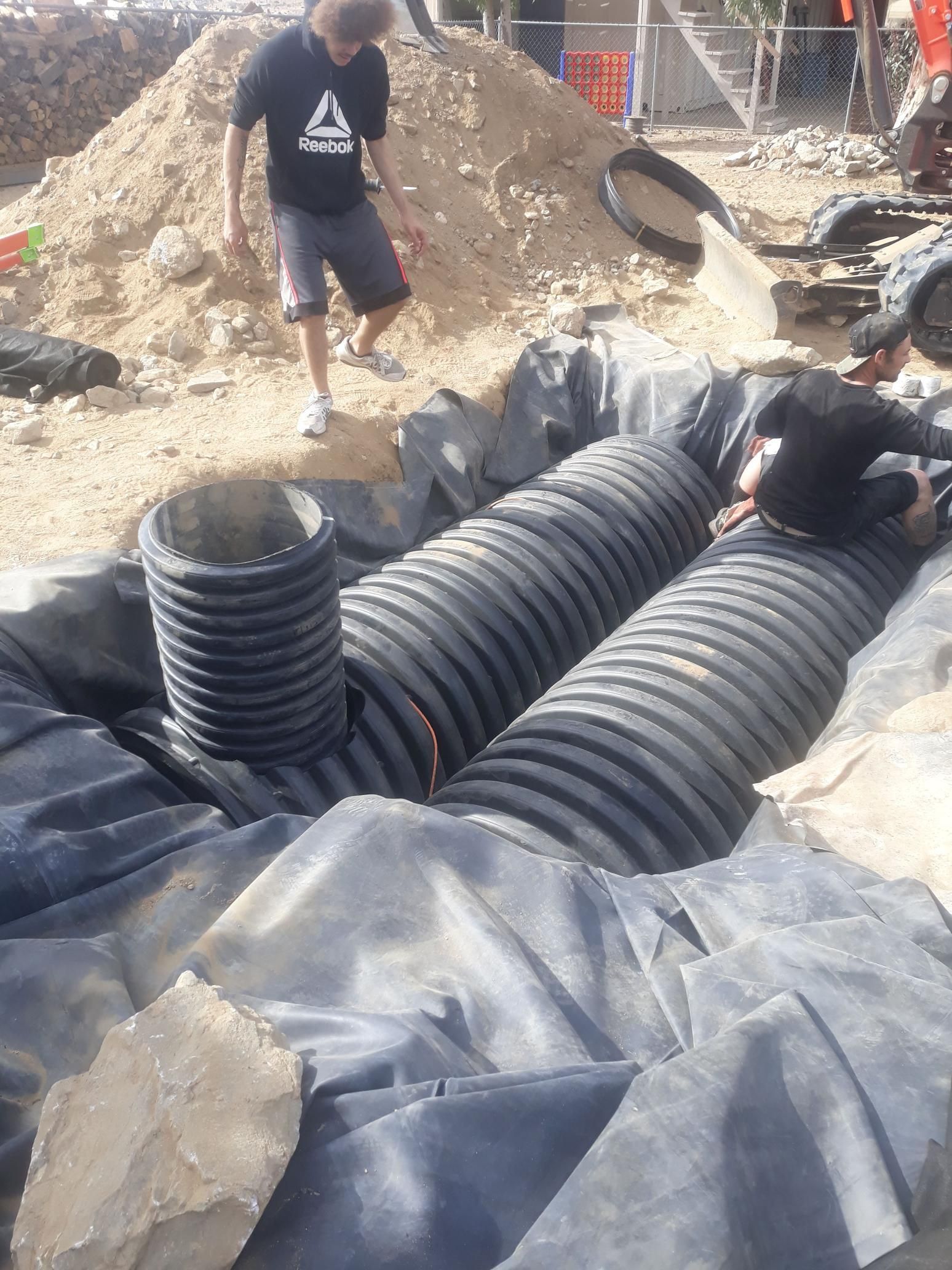 A man wearing a reebok shirt is pouring sand into a drainage system.