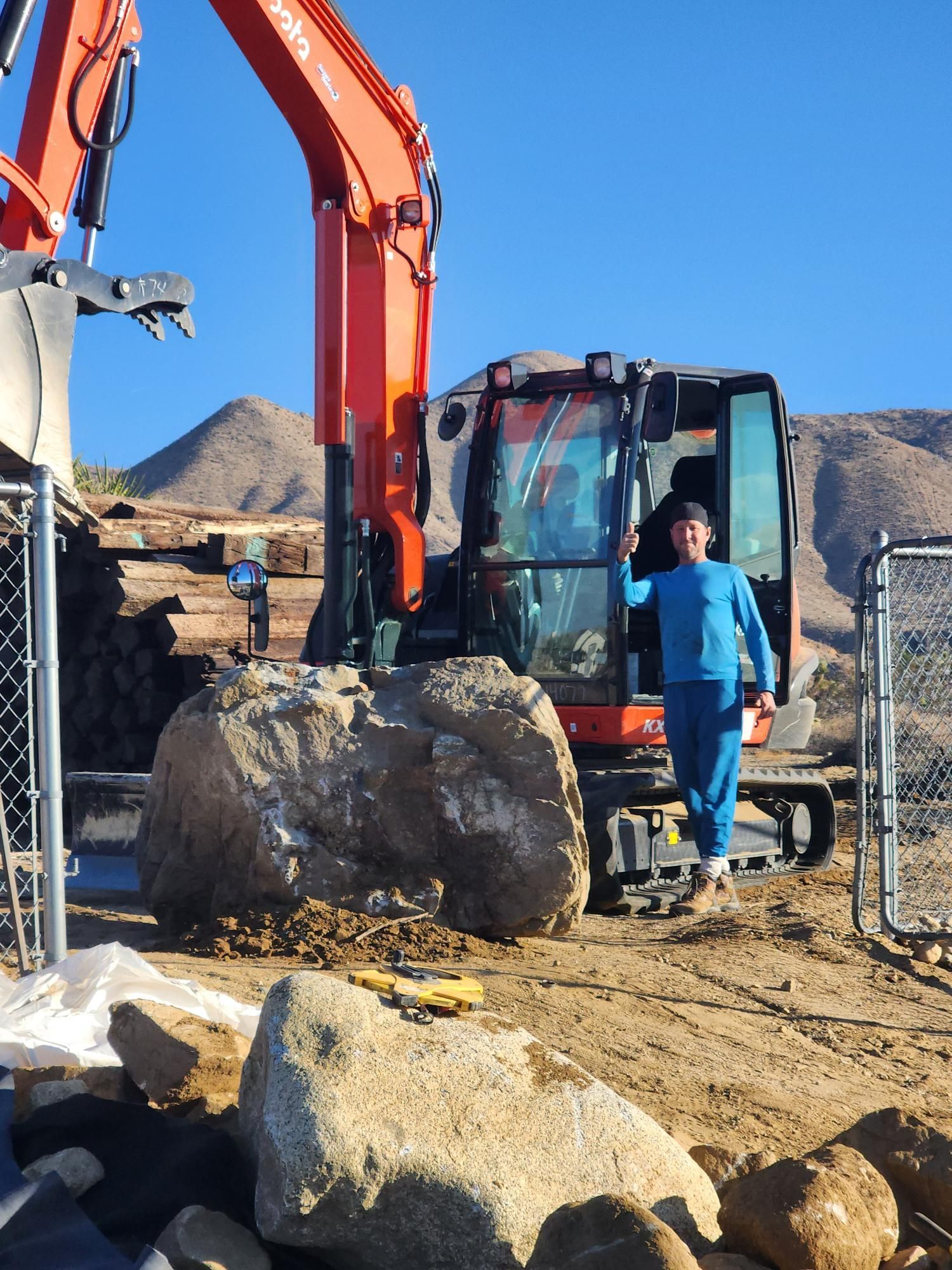 A man is standing in front of an excavator with a large rock in front of it