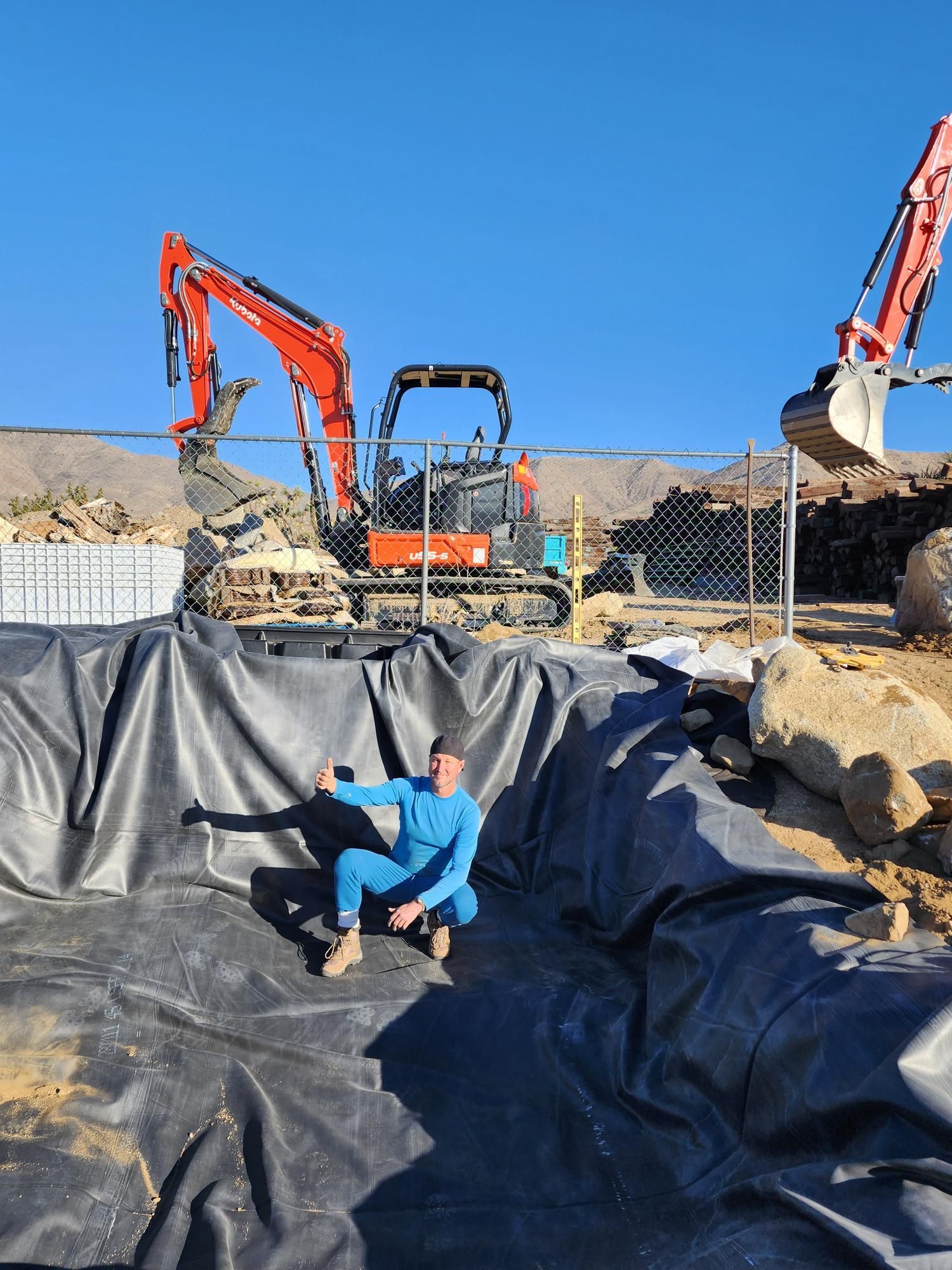 A man is kneeling on a black tarp in front of a large excavator.