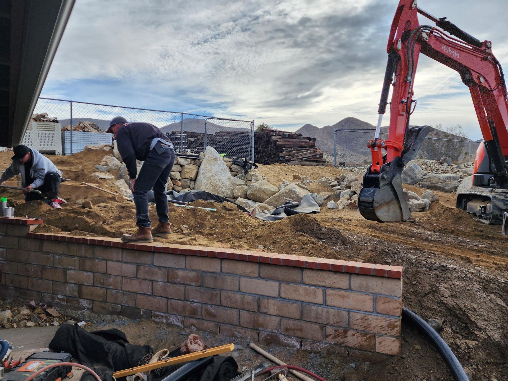 Two men are working on a brick wall next to an excavator.