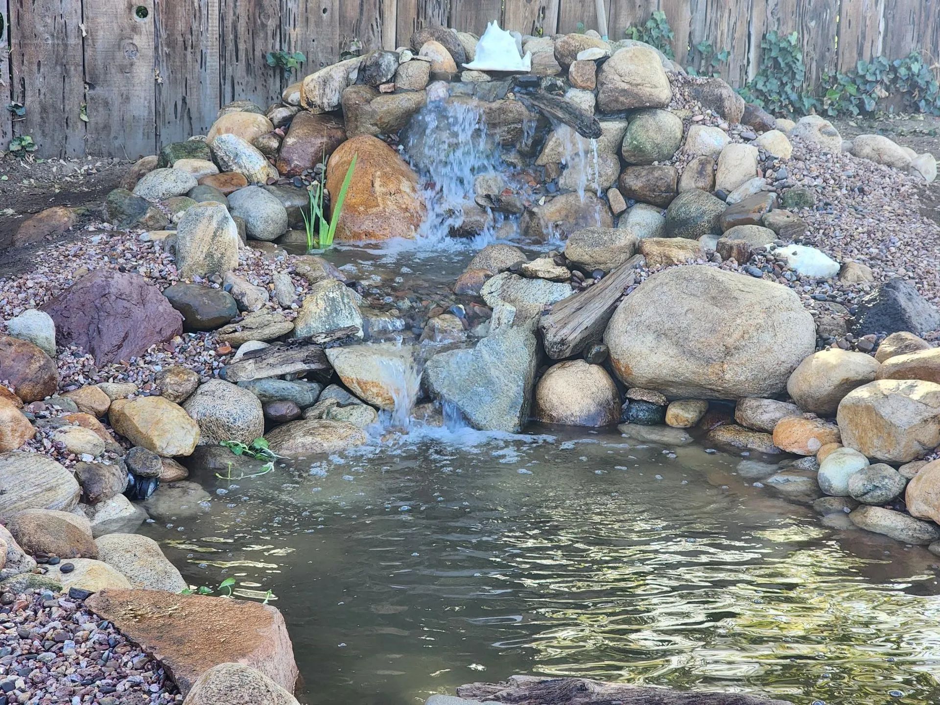 Rock waterfall flowing into a small pond, surrounded by stacked stones and a wooden fence