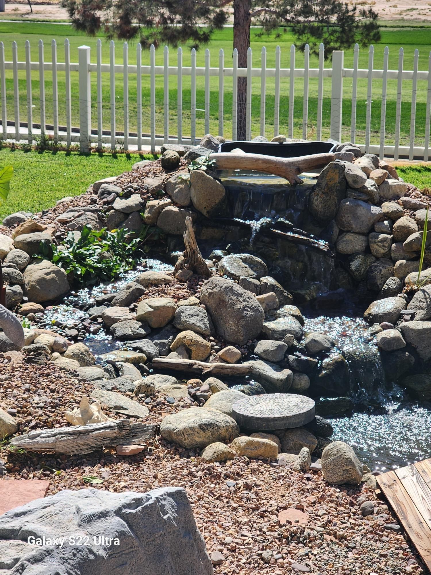 A waterfall is surrounded by rocks and a white fence.
