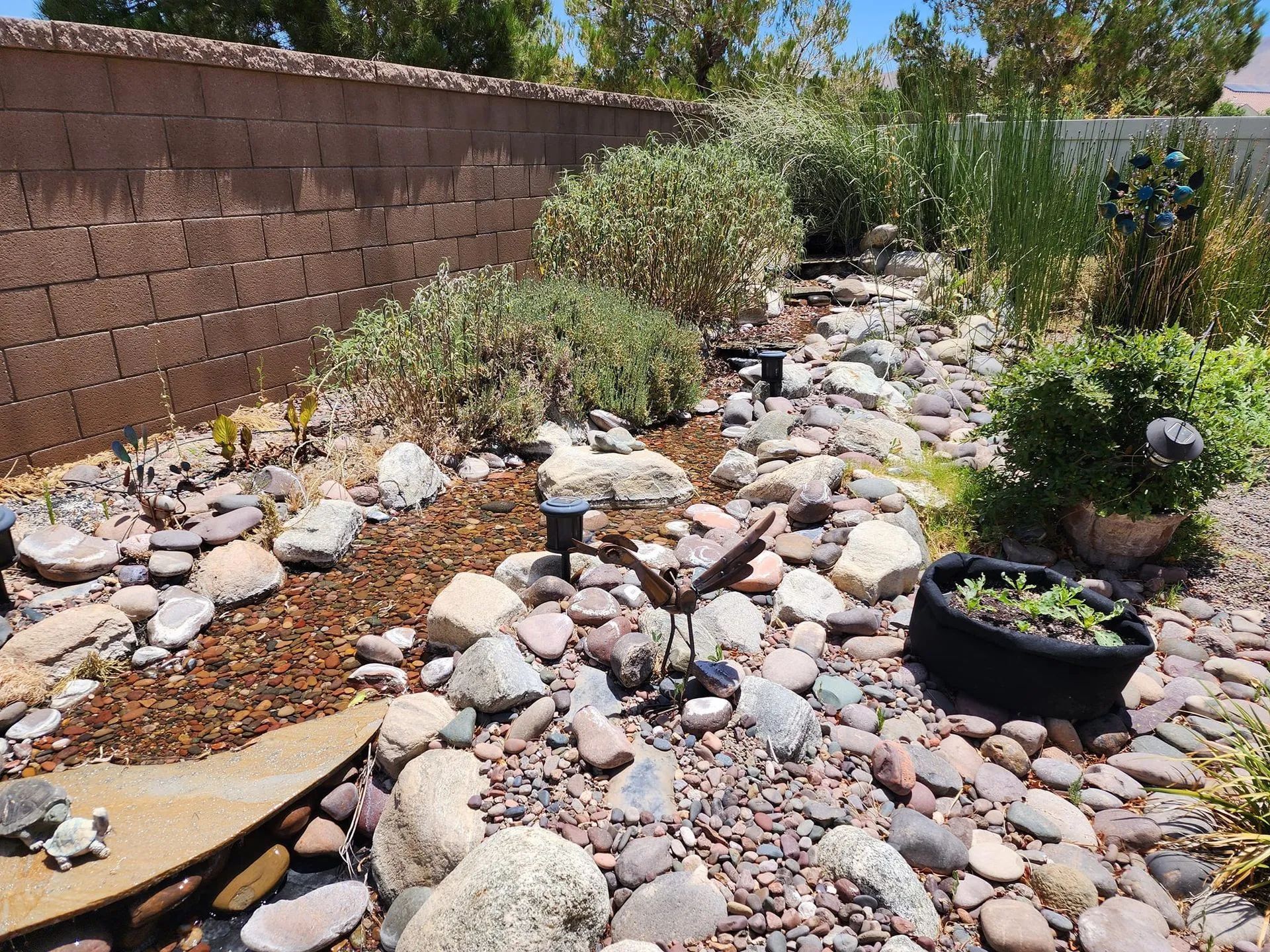 A dry creek bed landscaping design featuring various river stones, gravel, shrubs, and a small black planter.