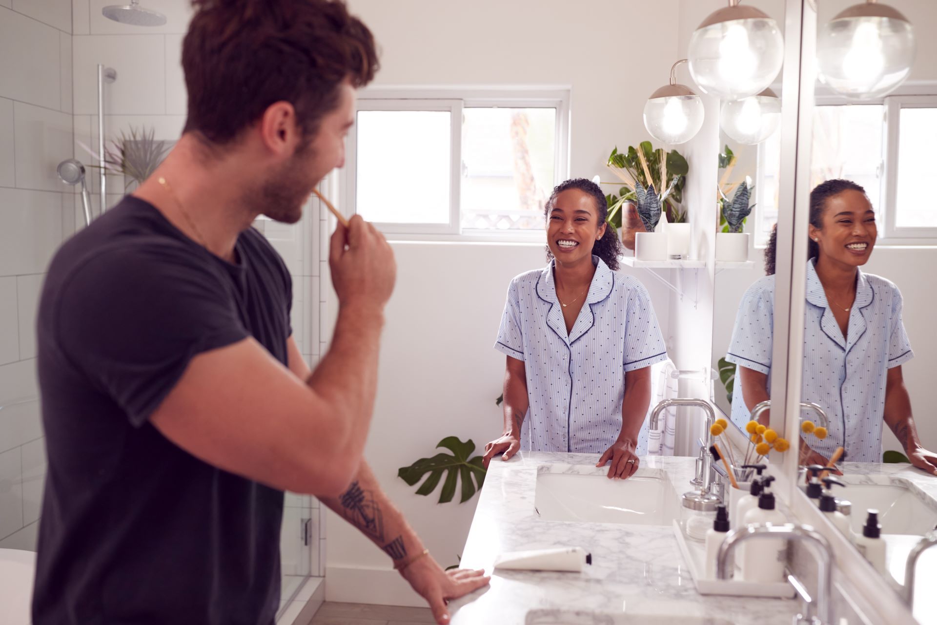 Man brushing teeth, woman smiling at him in a bathroom with plants and mirror.