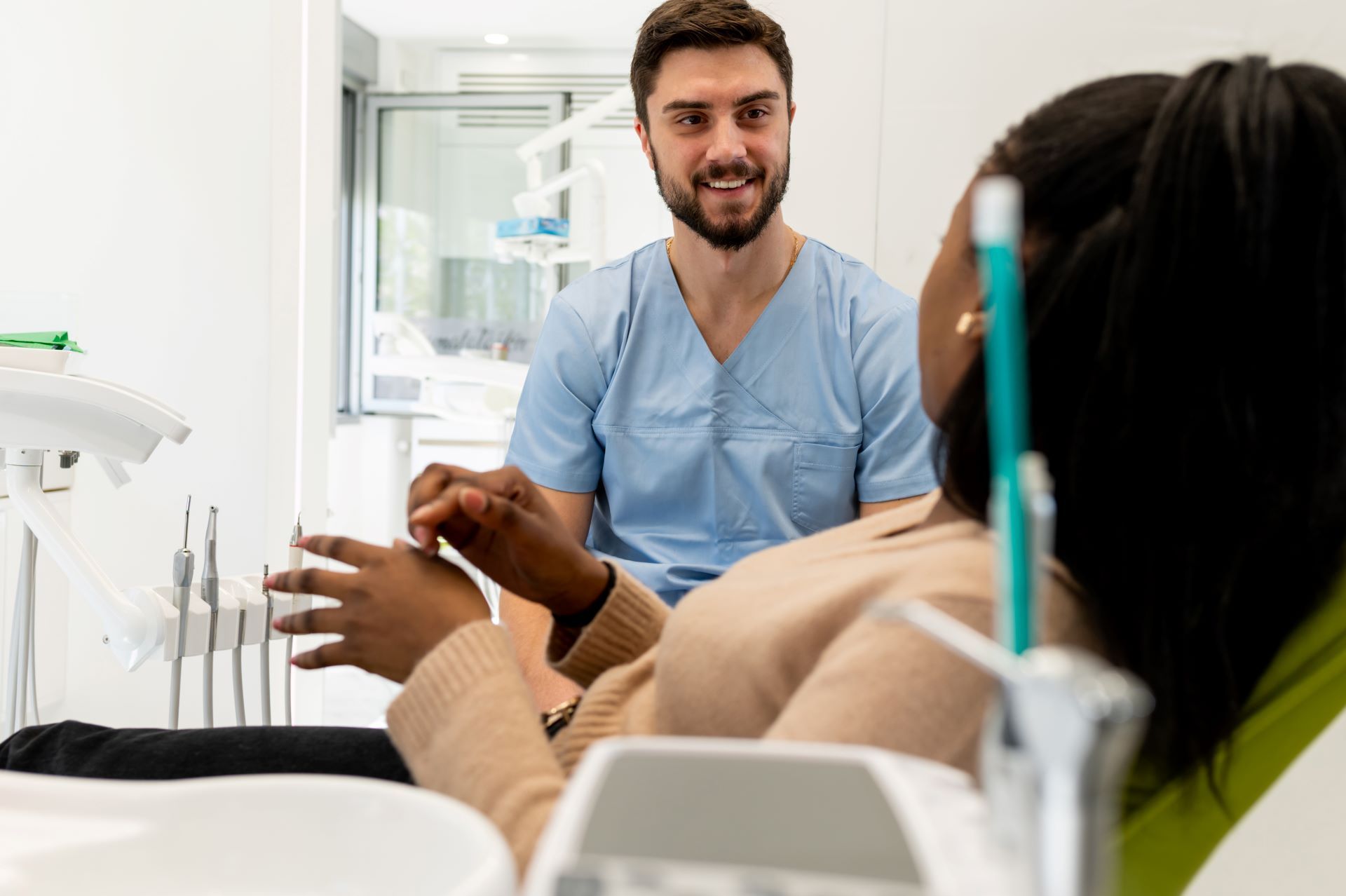 Dentist in blue scrubs smiles at patient in dental chair, modern office setting.