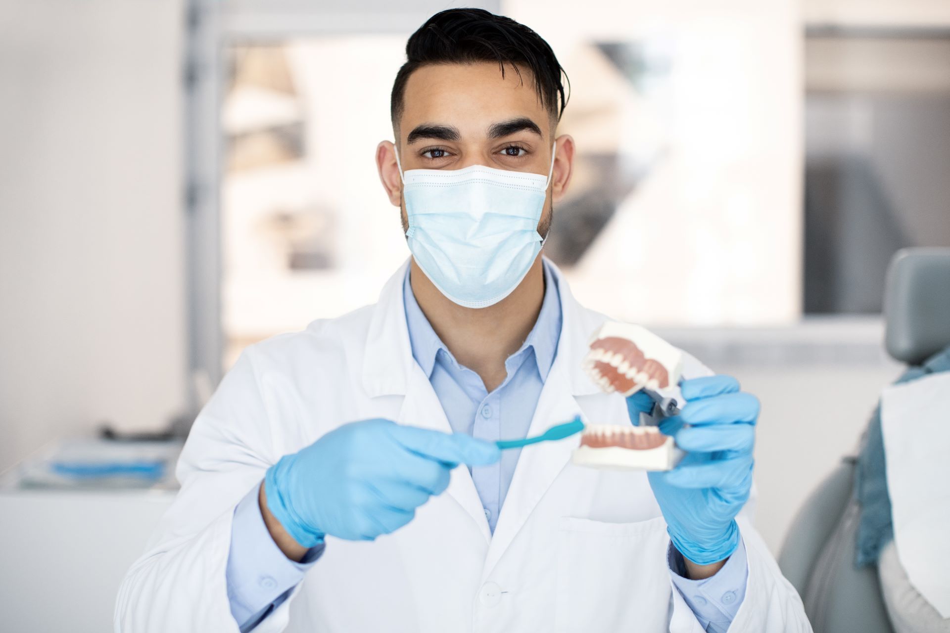 A dentist wearing a mask and gloves is brushing a model of teeth with a toothbrush.