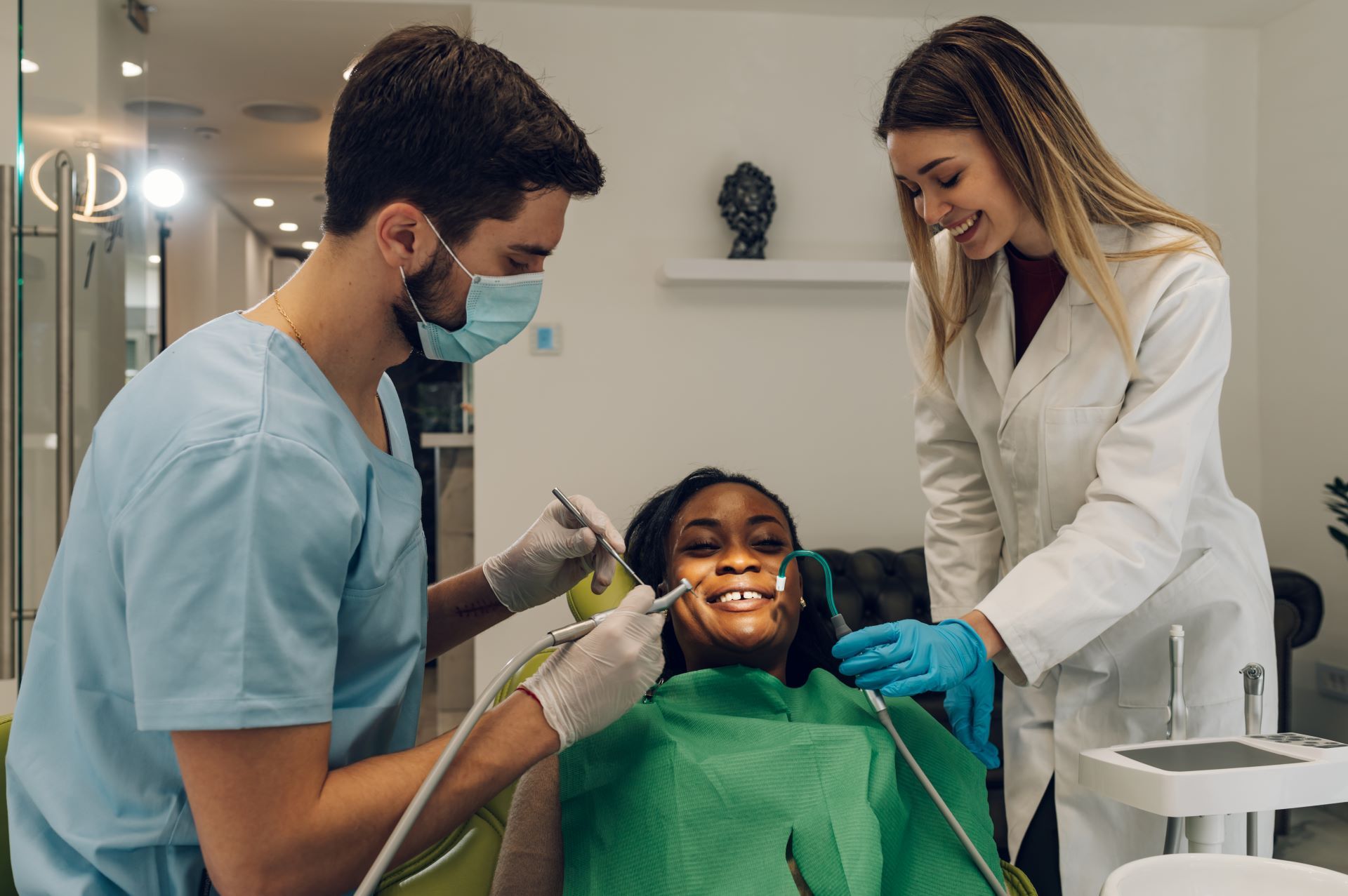 Dentist examining a Black woman's teeth while an assistant observes in a dental office.
