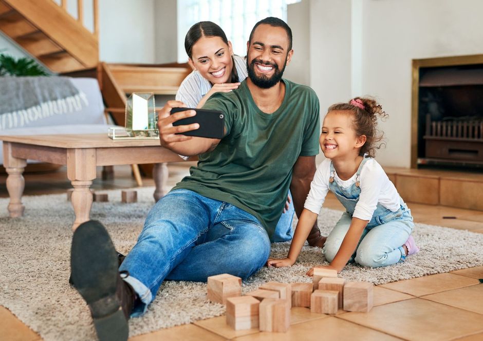 Family with mother, father and daughter all smiling while taking a selfie. Wooden toy blocks on the floor.