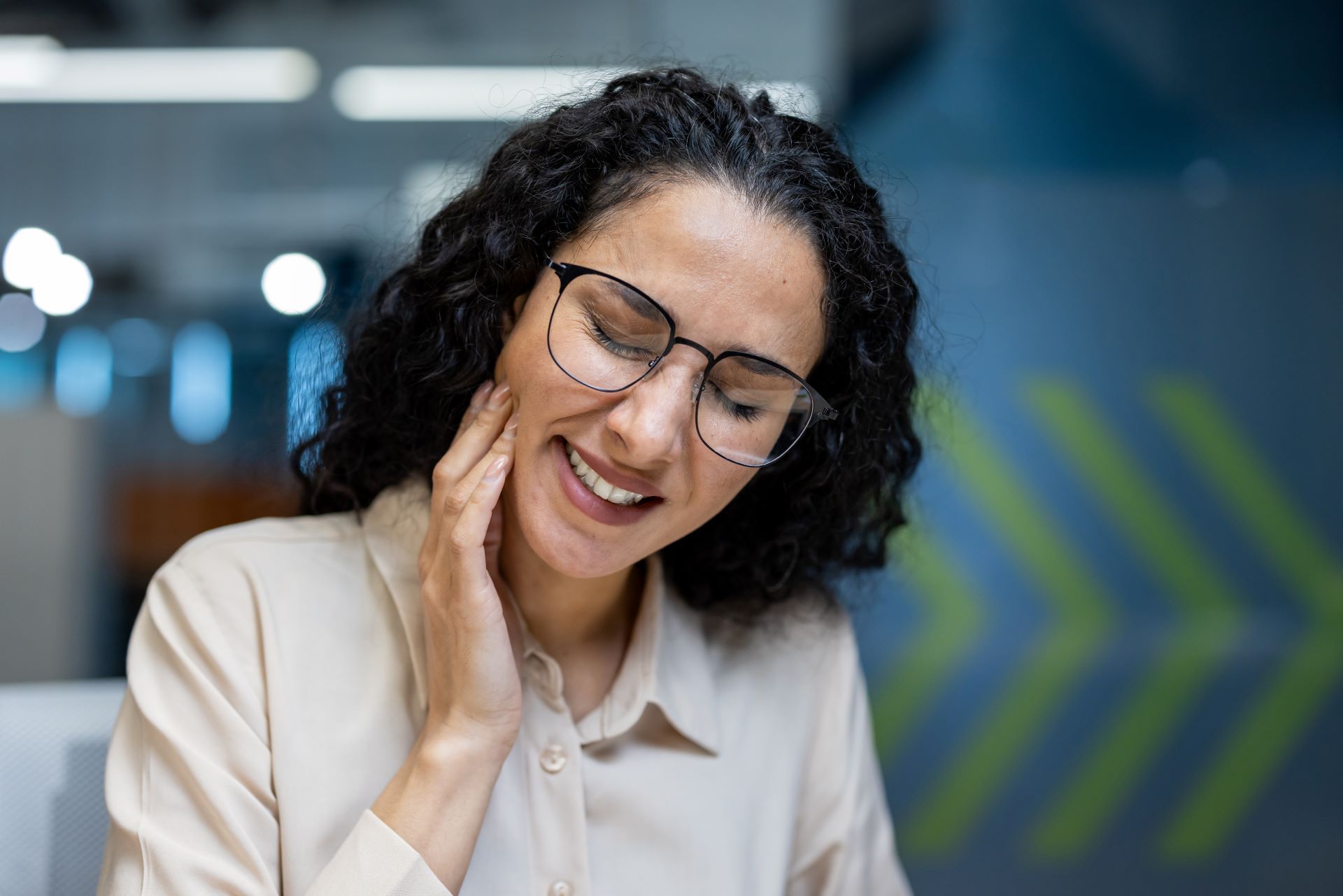 Woman with glasses, curly hair, holding her jaw, appears to be in pain. Office setting.