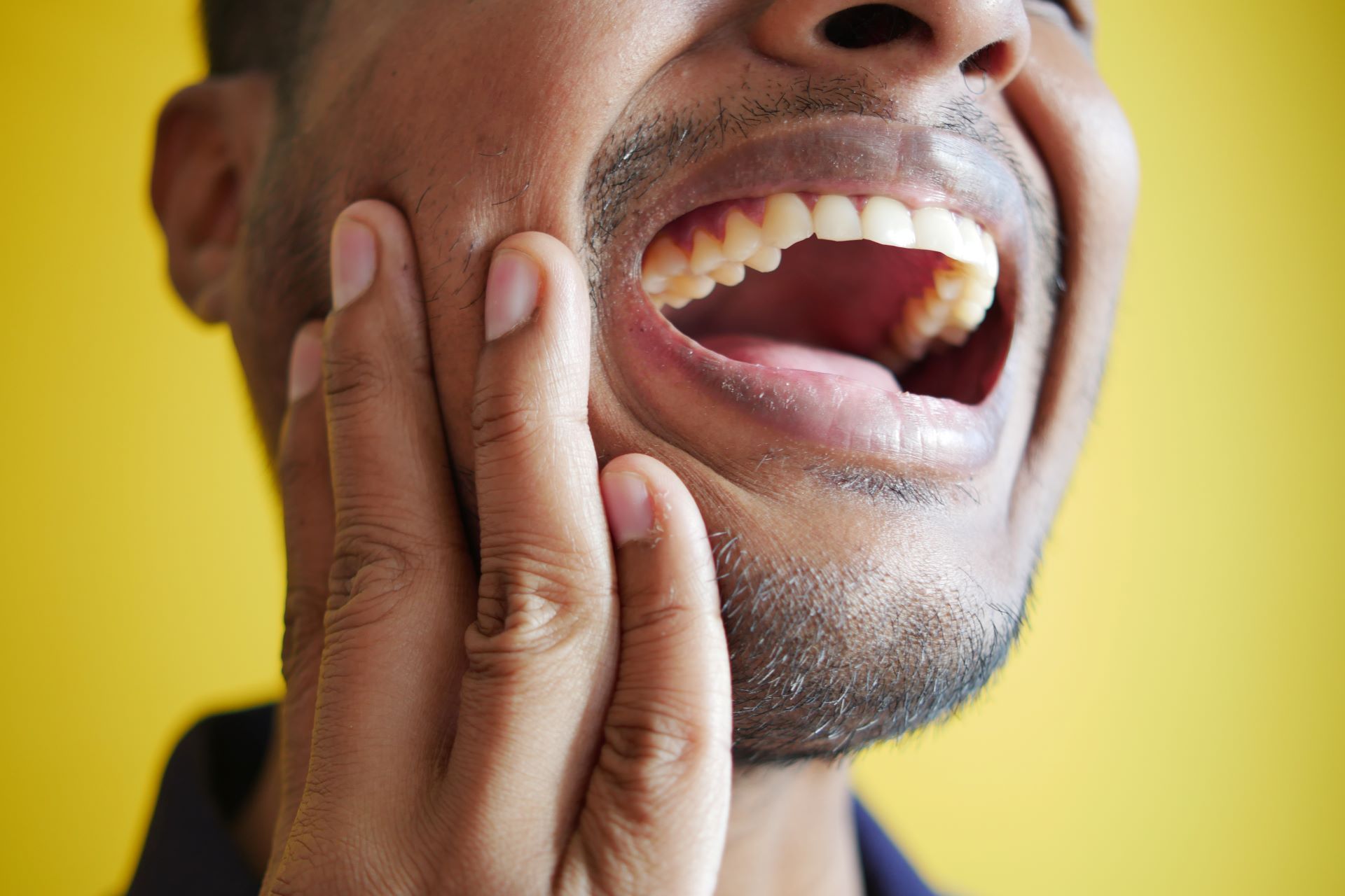 Man holding his cheek and open mouth, appearing to have tooth pain against a yellow background