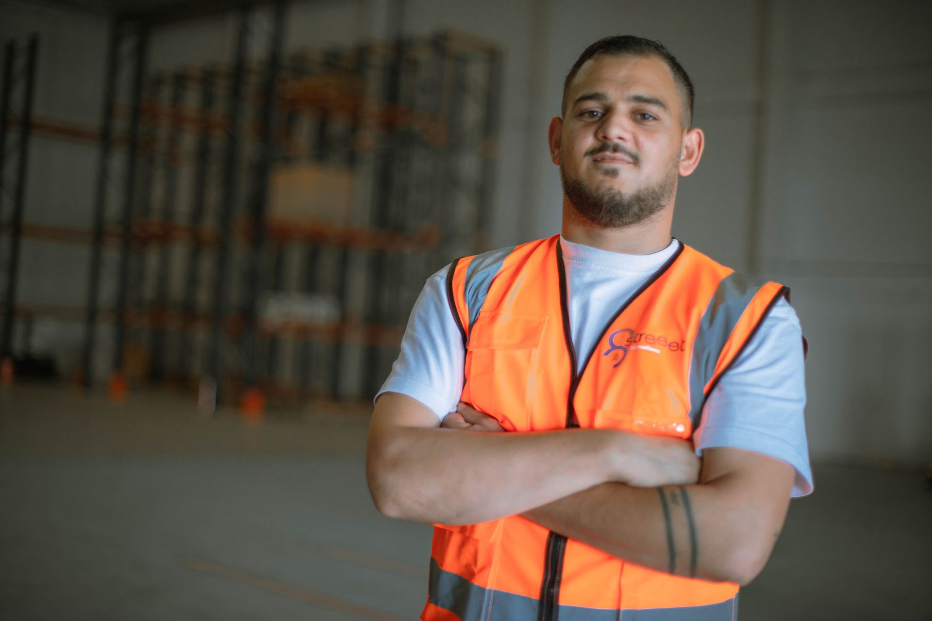 Un homme portant un gilet orange se tient debout dans un entrepôt, les bras croisés.