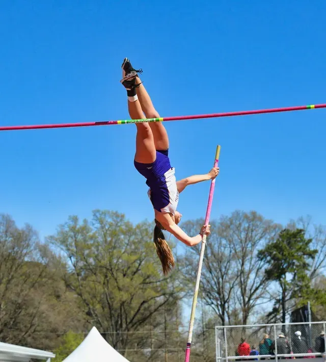 Pole vaulter upside down, clearing a bar, blue sky background.