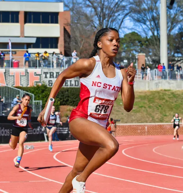 Track athlete in red and white NC State uniform sprints on track, holding a baton.