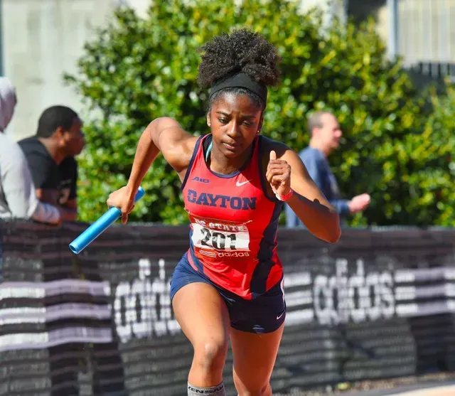 Track athlete with baton, wearing Dayton jersey, running outdoors.