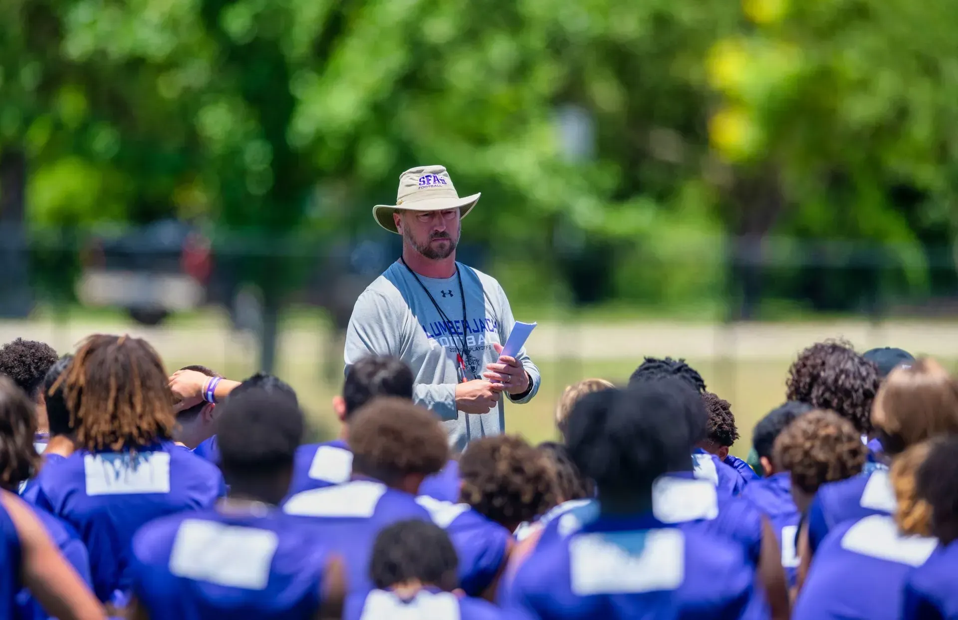 Football coach in a hat addressing players in blue jerseys on a sunny field.