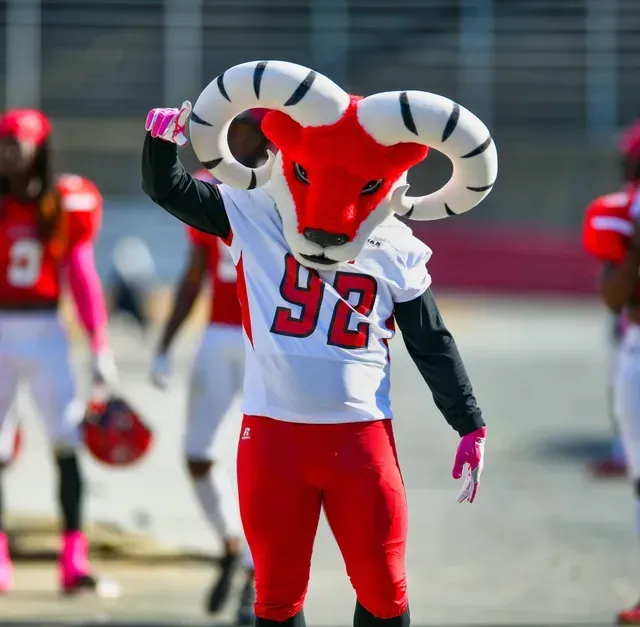 A ram mascot wearing a football jersey and red pants stands on a field.