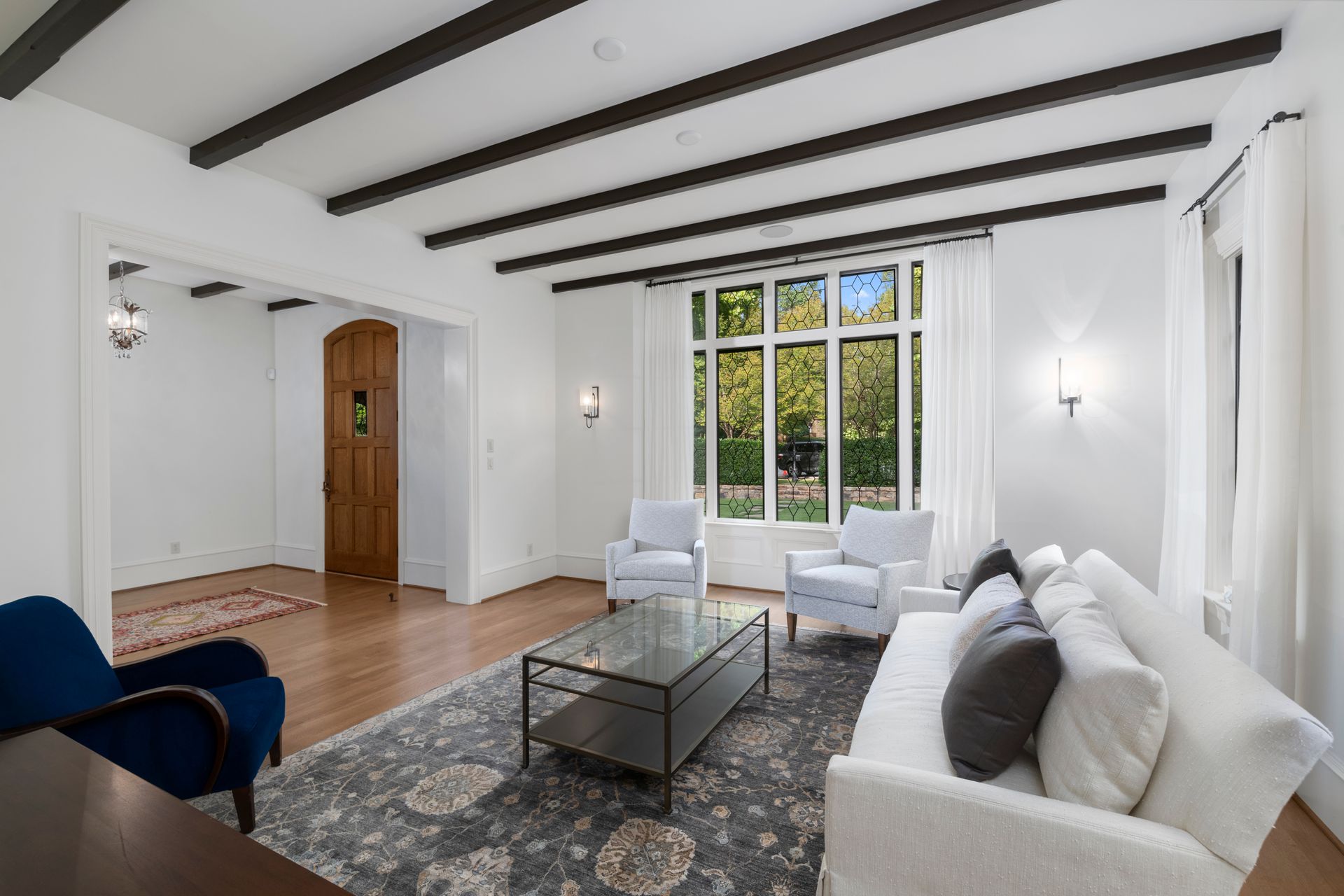 Bright living room with white sofa, blue chair, glass coffee table, exposed beams, and large windows.