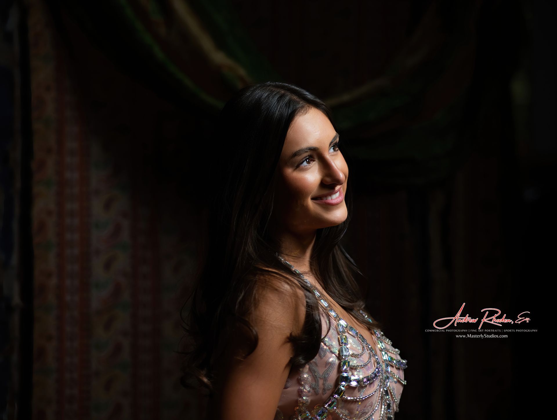 Woman smiles, in a shimmery gown, looking upward, against a dark backdrop with drapery.