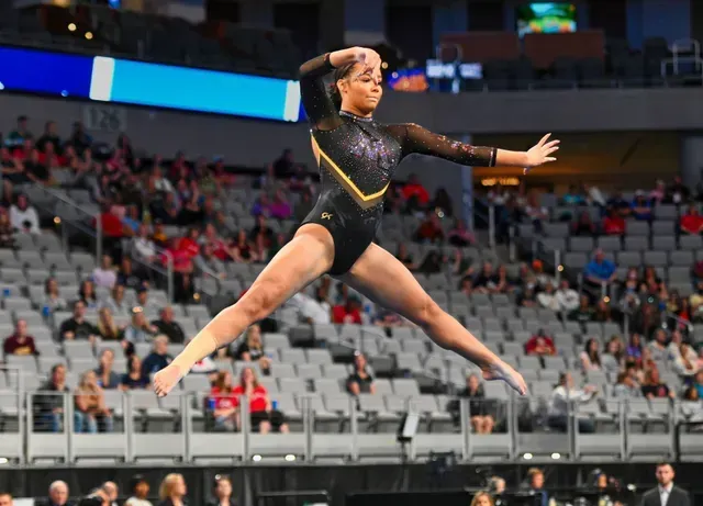 Gymnast in a black leotard performs a leap on a floor exercise, arena background.