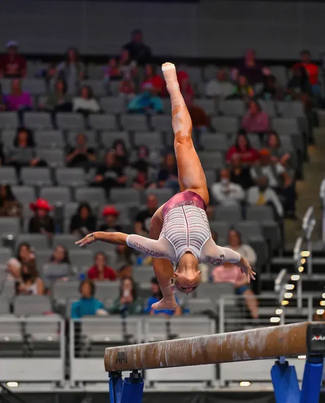 Gymnast performs a backflip on a balance beam in an arena with spectators.