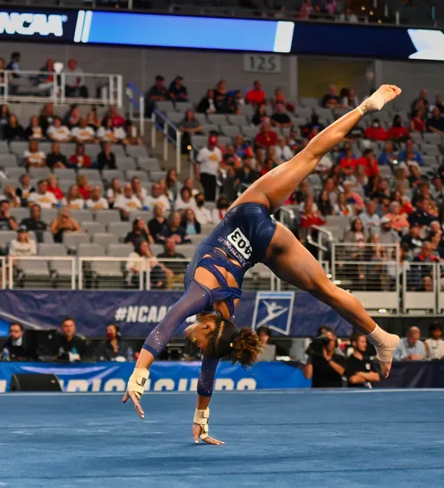 Gymnast performing a handstand with a leg split on a blue mat, in a competition setting.