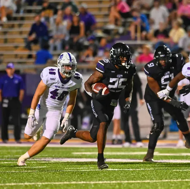 Football player in black uniform runs with ball, pursued by player in white uniform on green field.
