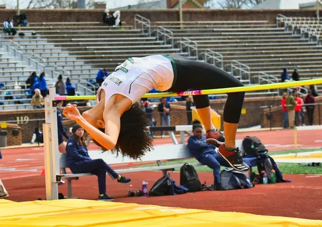 High jumper arches over bar on track; athlete in white top and black leggings, stadium in background.