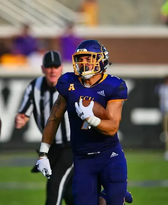 Football player in purple uniform running with ball on a field. A referee is in the background.