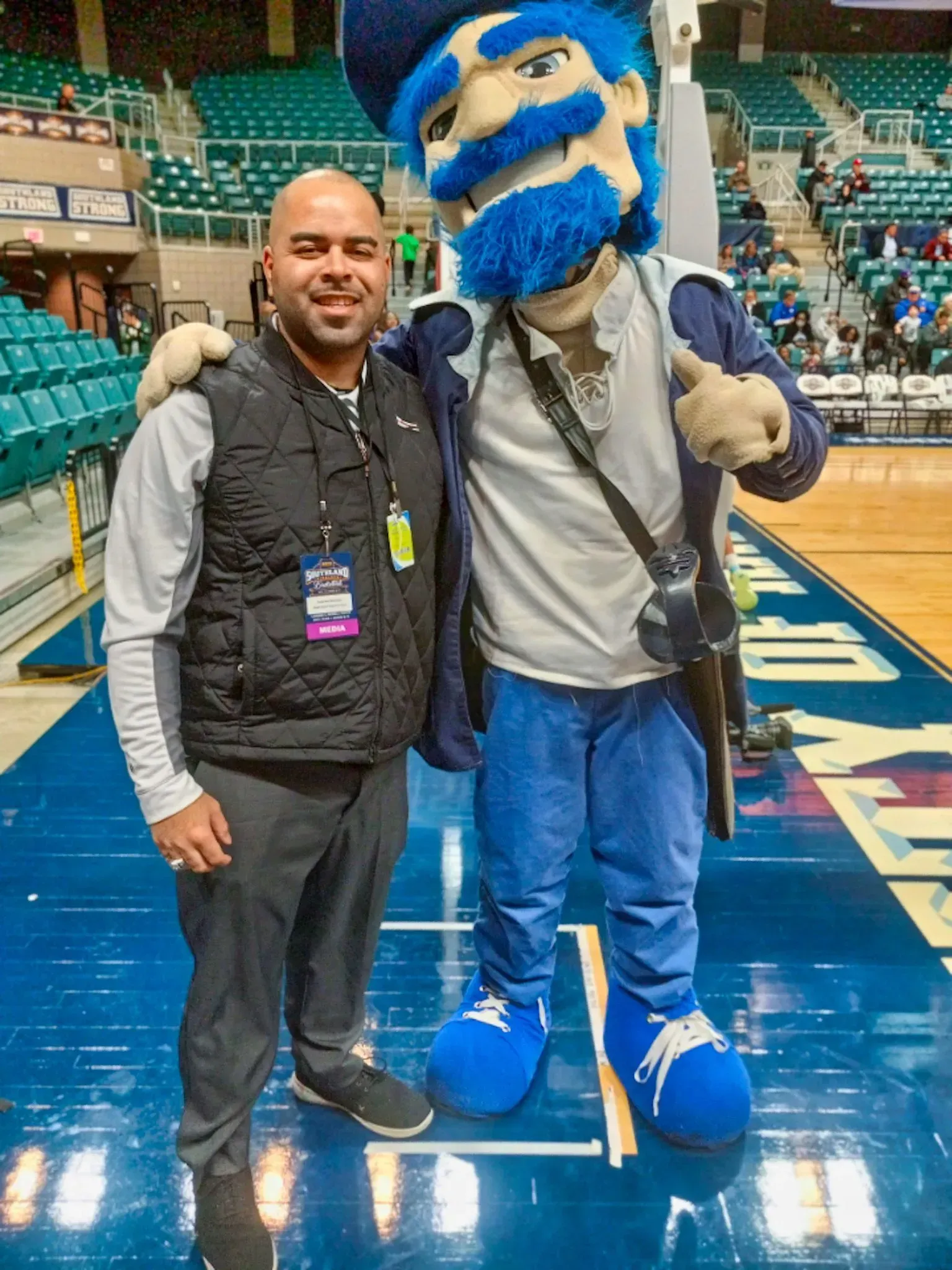 Man with arm around university mascot on basketball court. Mascot is in blue and white.
