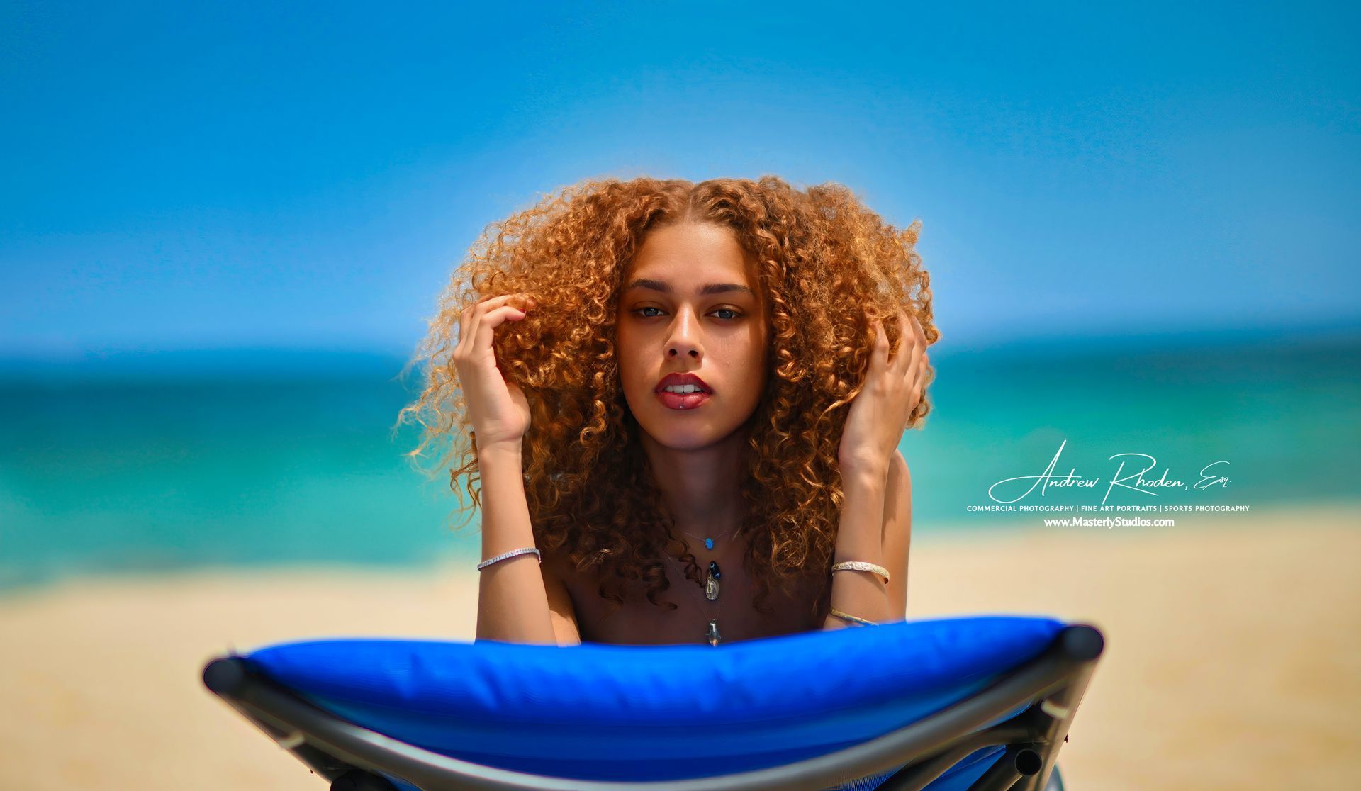 Woman with curly hair on a beach, holding her hair, looking towards the camera, blue ocean background.
