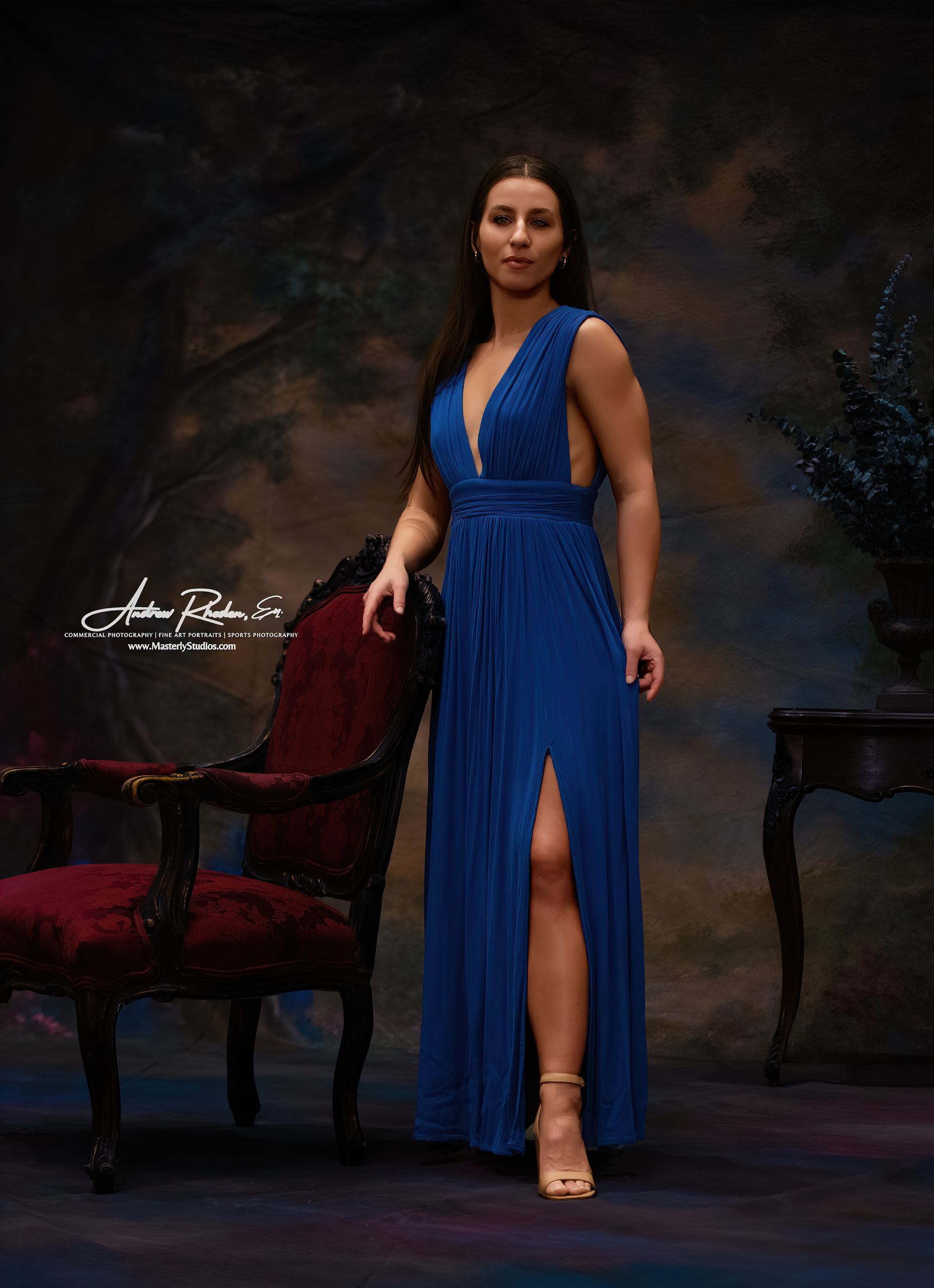 Woman in a blue gown standing beside a red chair against a dark studio backdrop.