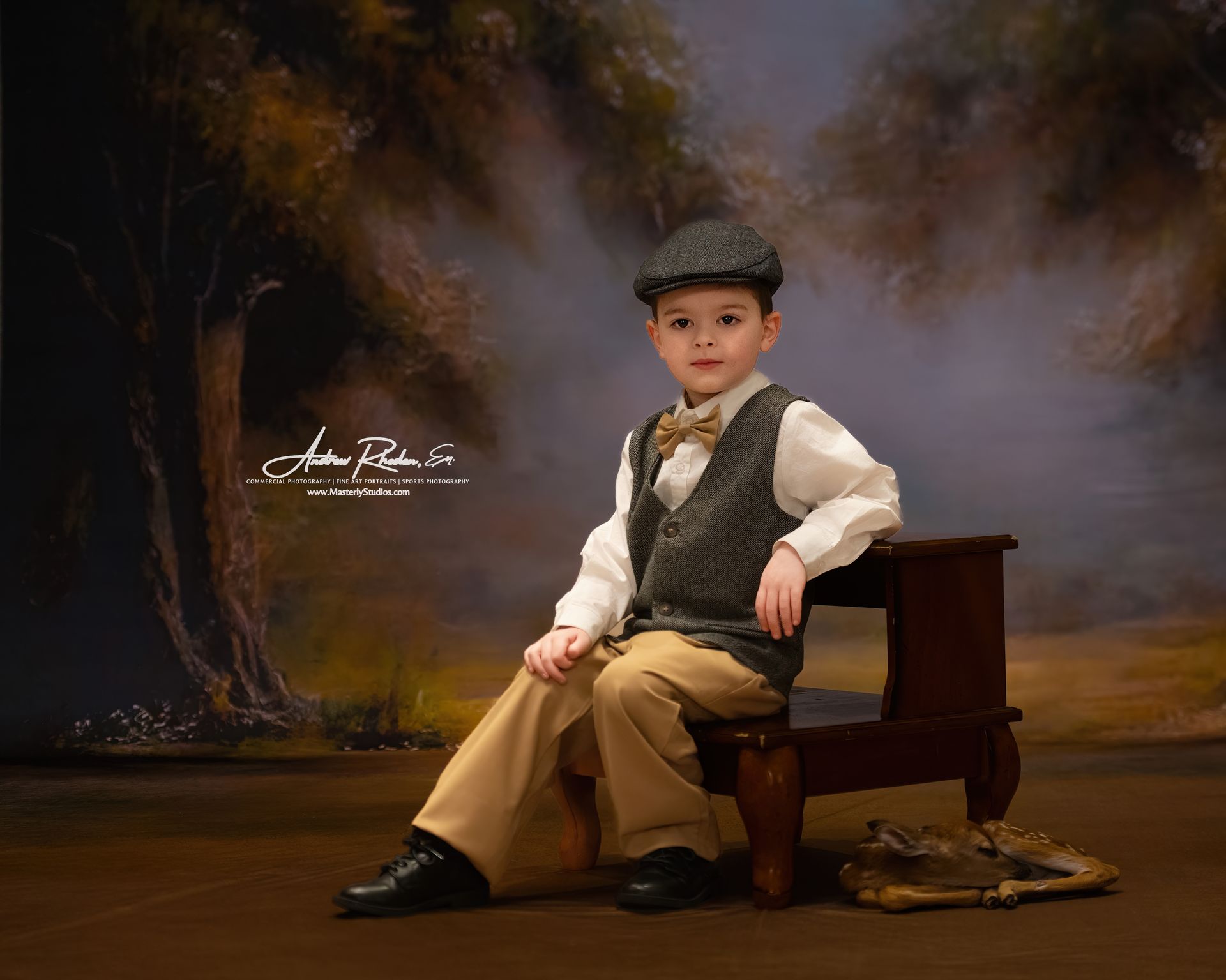 Young child in tweed vest and cap sits on a small stool against a blurred forest backdrop.