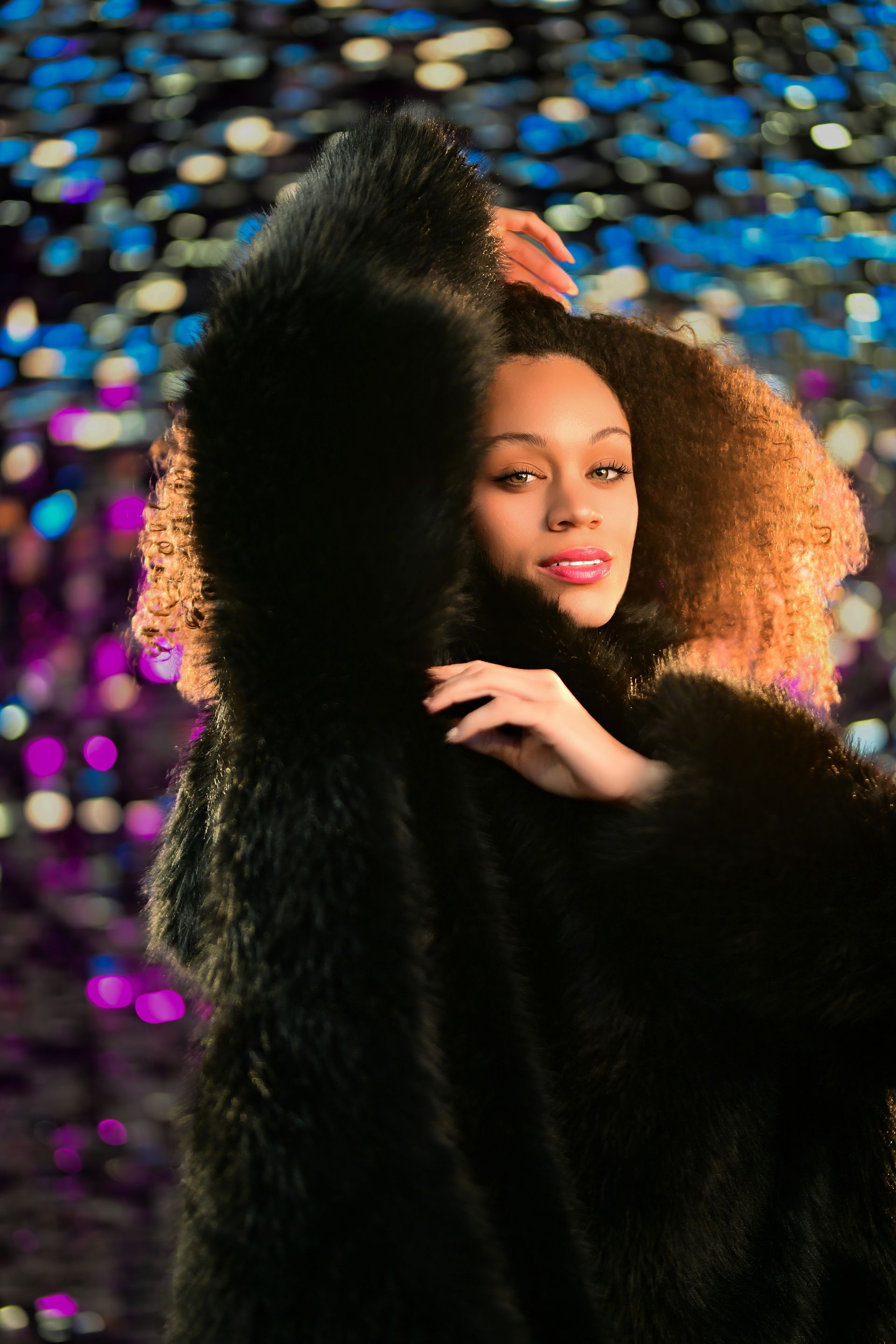 Person in a black fur coat posing against a colorful blurred lights background.