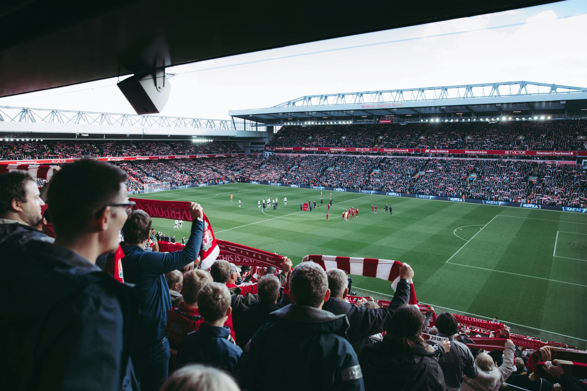Fans cheer at a packed soccer stadium, waving red and white scarves, watching a game on a green field.
